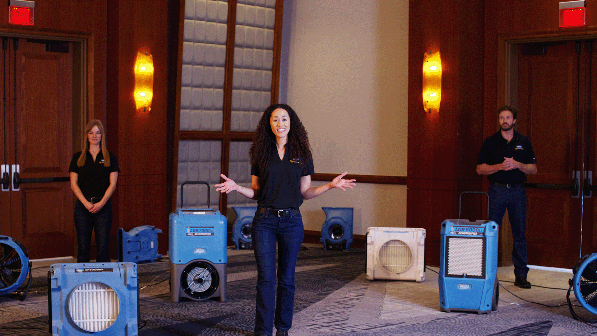 Three people standing in a room wearing R2R branded black shirts and jeans. They are surrounded by Legend Brands water mitigation equipment. Including dehumidifiers, airmovers, and fans.